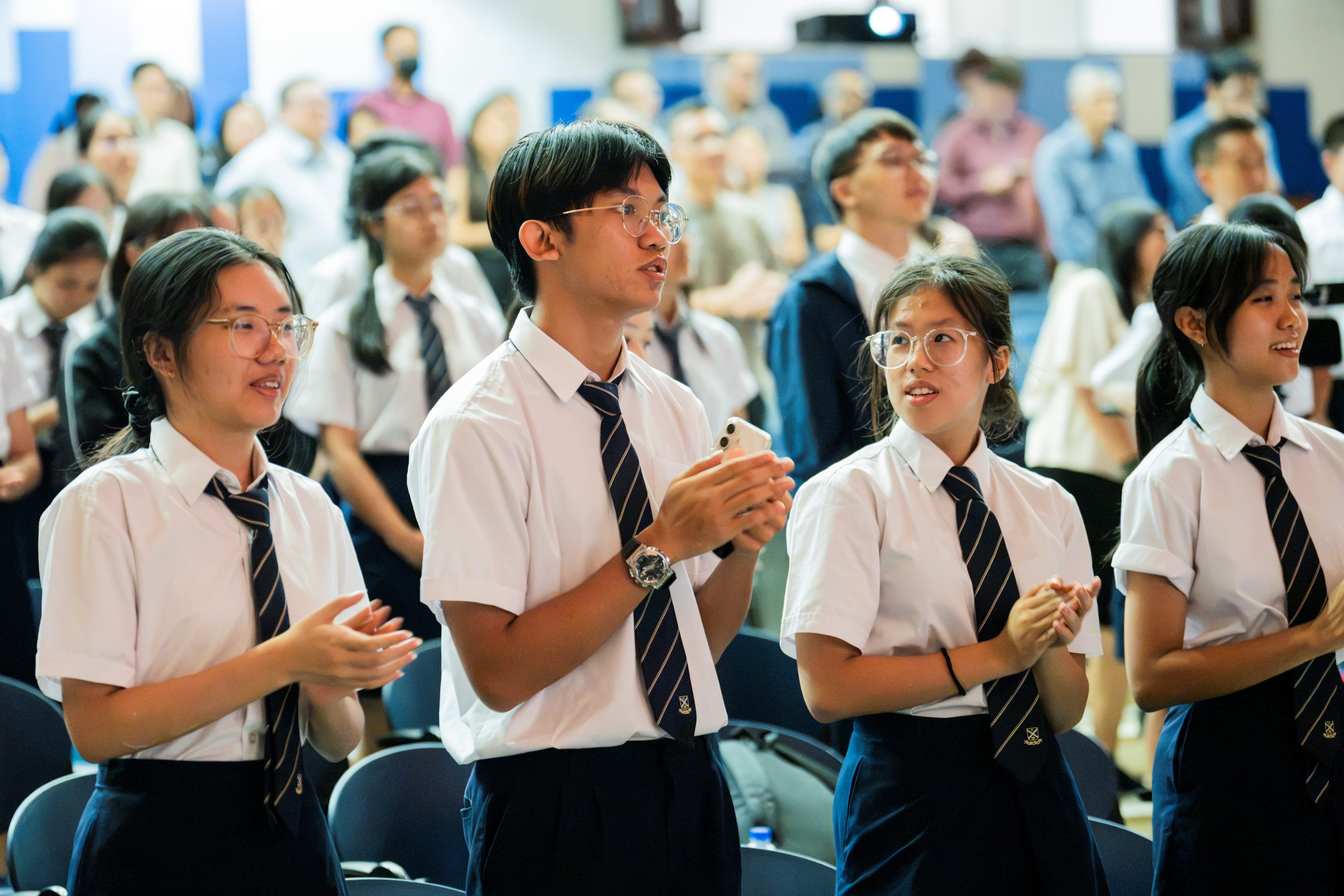 Students in school uniform standing and applauding during an event in an auditorium.